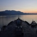 The image shows the bow of a large naval or commercial vessel photographed from an elevated stern perspective during twilight or early dawn. A tugboat with illuminated lights is positioned alongside the starboard side of the main vessel, likely assisting with maneuvering. A mountainous coastline with a small settlement is visible in the background across calm waters.