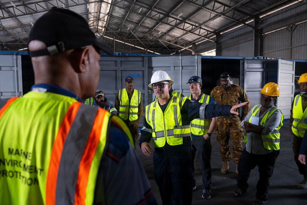 A group of workers and personnel are gathered inside a large industrial warehouse or hangar facility. The group includes individuals wearing high-visibility safety vests, hard hats, and military camouflage uniforms, suggesting a multi-agency or joint operational briefing. Shipping containers are visible in the background, indicating a logistics or port-related environment.