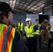 A group of workers and personnel are gathered inside a large industrial warehouse or hangar facility. The group includes individuals wearing high-visibility safety vests, hard hats, and military camouflage uniforms, suggesting a multi-agency or joint operational briefing. Shipping containers are visible in the background, indicating a logistics or port-related environment.