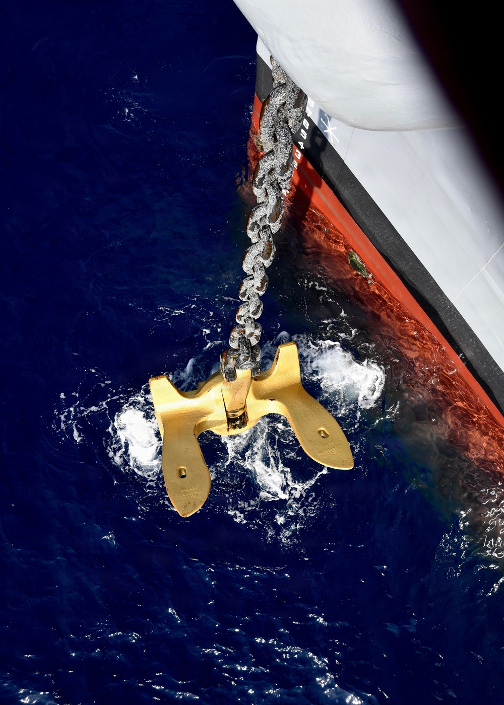 An aerial/overhead view of a large ship's anchor being deployed or retrieved at the waterline. The anchor, finished in bright gold/yellow paint, is suspended by a heavy metal chain extending from the ship's hull. The ship's hull displays the characteristic red anti-fouling paint below the waterline and white above, with churning white water surrounding the anchor.