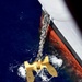 An aerial/overhead view of a large ship's anchor being deployed or retrieved at the waterline. The anchor, finished in bright gold/yellow paint, is suspended by a heavy metal chain extending from the ship's hull. The ship's hull displays the characteristic red anti-fouling paint below the waterline and white above, with churning white water surrounding the anchor.