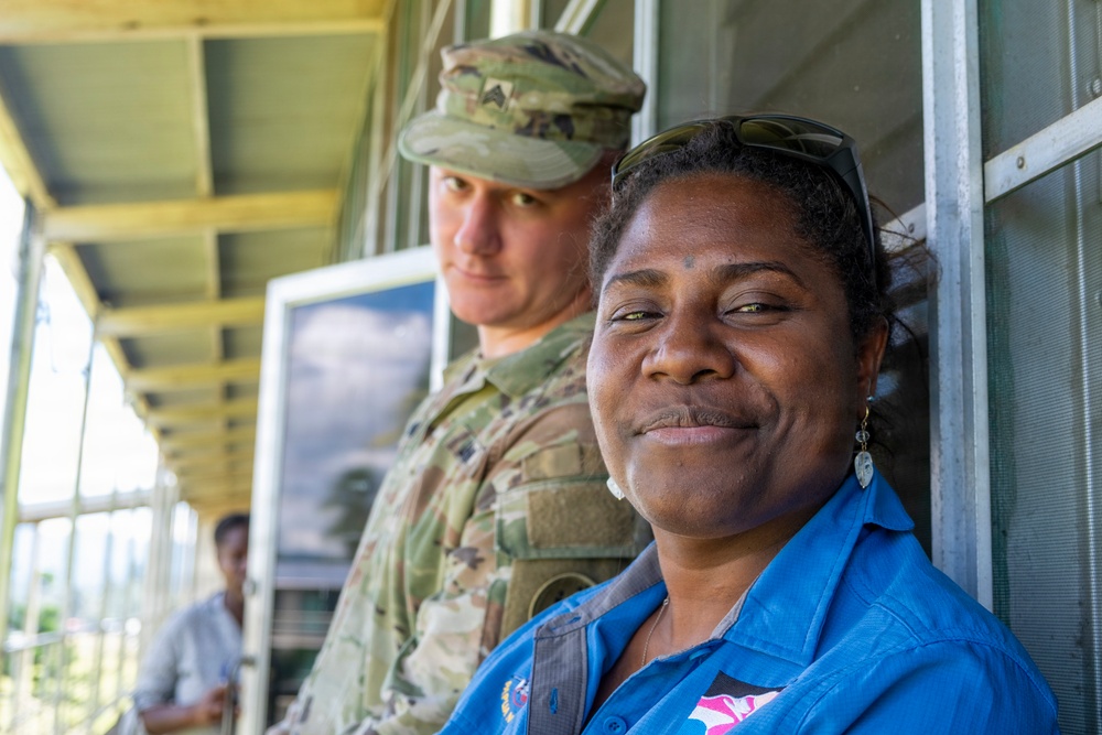 A woman in a blue shirt with sunglasses on her head smiles warmly at the camera while standing on a covered porch or veranda. Behind her stands a male soldier in multicam/OCP camouflage uniform and patrol cap. A third person is partially visible in the background on the left.