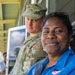 A woman in a blue shirt with sunglasses on her head smiles warmly at the camera while standing on a covered porch or veranda. Behind her stands a male soldier in multicam/OCP camouflage uniform and patrol cap. A third person is partially visible in the background on the left.