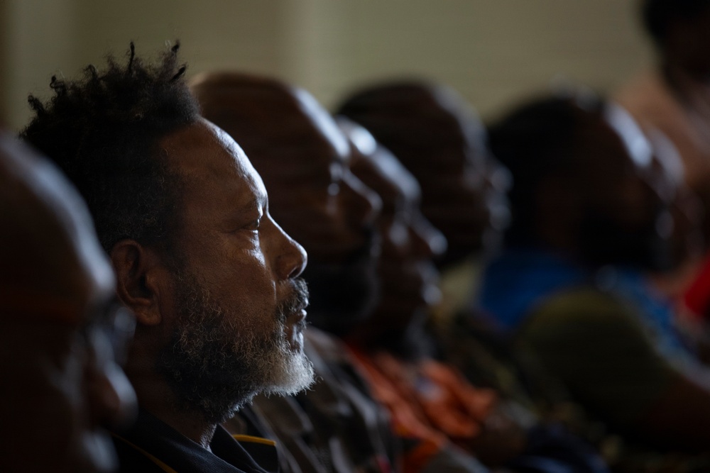 A group of people are seated in rows, appearing to be attending a gathering, meeting, or service. The foreground subject is a middle-aged man with a gray beard and natural hair, photographed in sharp profile with dramatic side lighting. The background figures are progressively out of focus, creating a shallow depth-of-field effect.