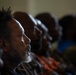 A group of people are seated in rows, appearing to be attending a gathering, meeting, or service. The foreground subject is a middle-aged man with a gray beard and natural hair, photographed in sharp profile with dramatic side lighting. The background figures are progressively out of focus, creating a shallow depth-of-field effect.