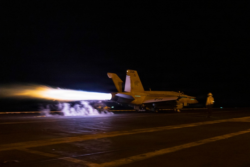 A military jet aircraft, marked '306', is captured during a night catapult launch from what appears to be an aircraft carrier flight deck. The aircraft's afterburners are fully engaged, producing an intense elongated flame trail visible to the left of the aircraft. The long-exposure photograph captures the dramatic light streaks from the engine exhaust against the dark night sky.
