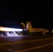 A military jet aircraft, marked '306', is captured during a night catapult launch from what appears to be an aircraft carrier flight deck. The aircraft's afterburners are fully engaged, producing an intense elongated flame trail visible to the left of the aircraft. The long-exposure photograph captures the dramatic light streaks from the engine exhaust against the dark night sky.