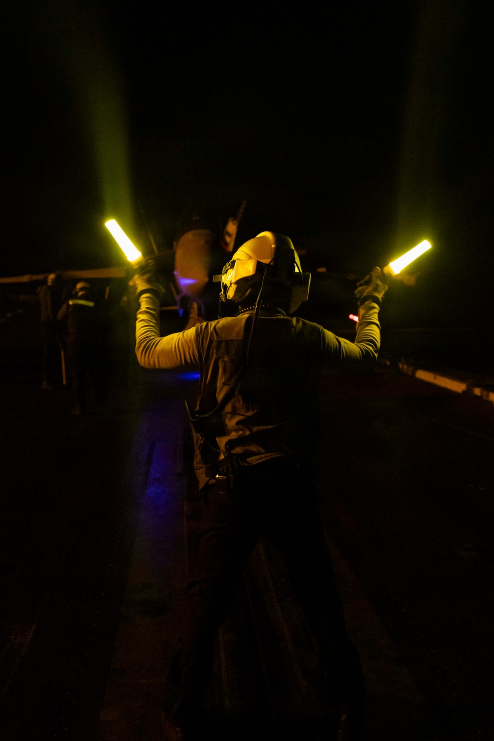A person wearing a helmet with night vision or protective gear stands on what appears to be a dark flight deck or tarmac at night, holding two illuminated yellow/amber light wands raised above their shoulders. The individual is wearing a dark vest over a long-sleeve yellow-green shirt and appears to be a flight deck crew member or aircraft handler. The scene is predominantly dark with the light wands serving as the primary light source illuminating the subject.