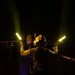A person wearing a helmet with night vision or protective gear stands on what appears to be a dark flight deck or tarmac at night, holding two illuminated yellow/amber light wands raised above their shoulders. The individual is wearing a dark vest over a long-sleeve yellow-green shirt and appears to be a flight deck crew member or aircraft handler. The scene is predominantly dark with the light wands serving as the primary light source illuminating the subject.