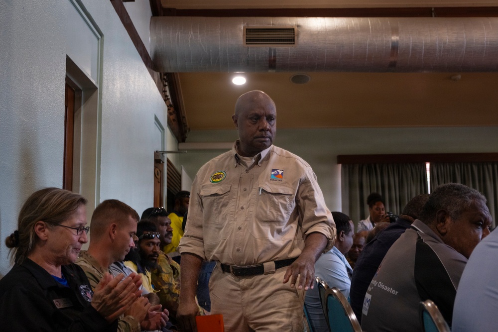 A man in a khaki uniform shirt with patches stands in the aisle of what appears to be an indoor meeting or community gathering. He is surrounded by seated attendees in a room with exposed ductwork on the ceiling. The atmosphere suggests a formal or semi-formal community event or briefing.