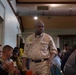 A man in a khaki uniform shirt with patches stands in the aisle of what appears to be an indoor meeting or community gathering. He is surrounded by seated attendees in a room with exposed ductwork on the ceiling. The atmosphere suggests a formal or semi-formal community event or briefing.