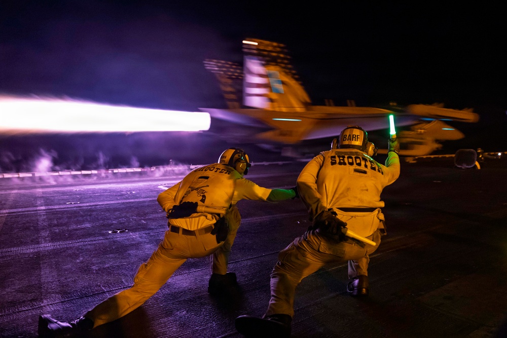 A nighttime aircraft carrier flight deck launch operation showing two catapult officers ('Shooters') signaling the launch of a military jet aircraft. The scene is dramatically lit by the aircraft's afterburner exhaust creating bright streaks of light against the dark sky. Motion blur on the aircraft indicates a high-speed catapult launch in progress.