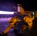 A nighttime aircraft carrier flight deck launch operation showing two catapult officers ('Shooters') signaling the launch of a military jet aircraft. The scene is dramatically lit by the aircraft's afterburner exhaust creating bright streaks of light against the dark sky. Motion blur on the aircraft indicates a high-speed catapult launch in progress.