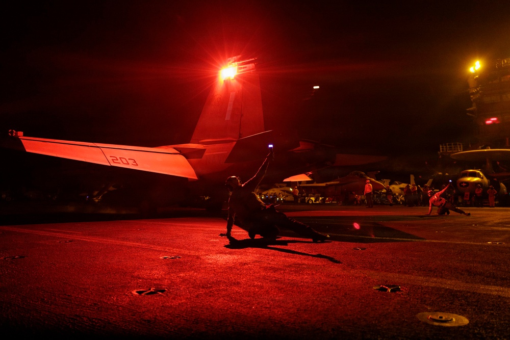 A nighttime flight deck operations scene aboard what appears to be a US Navy aircraft carrier. A deck crew member is visible in silhouette, raising a lighted wand as part of aircraft marshaling or launch signaling procedures. The scene is dramatically illuminated by a bright red anti-collision light mounted on the tail of a jet aircraft bearing the modex number '203'.