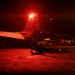 A nighttime flight deck operations scene aboard what appears to be a US Navy aircraft carrier. A deck crew member is visible in silhouette, raising a lighted wand as part of aircraft marshaling or launch signaling procedures. The scene is dramatically illuminated by a bright red anti-collision light mounted on the tail of a jet aircraft bearing the modex number '203'.