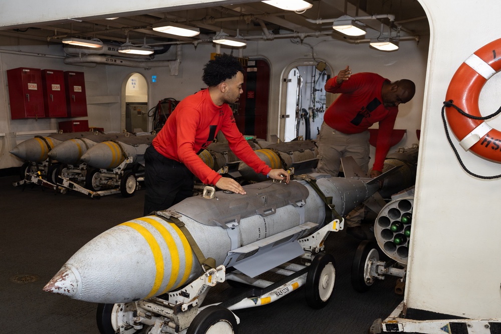 Two personnel wearing red shirts are handling munitions aboard what appears to be a naval vessel, likely an aircraft carrier's weapons handling area or hangar deck. The foreground shows a large bomb or ordnance on a wheeled transport dolly with distinctive yellow bands on its nose cone. Multiple additional munitions on dollies are visible in the background.
