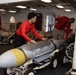 Two personnel wearing red shirts are handling munitions aboard what appears to be a naval vessel, likely an aircraft carrier's weapons handling area or hangar deck. The foreground shows a large bomb or ordnance on a wheeled transport dolly with distinctive yellow bands on its nose cone. Multiple additional munitions on dollies are visible in the background.