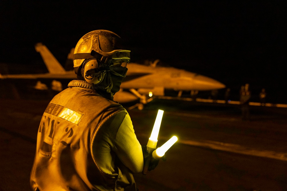 A flight deck crew member is conducting night operations on what appears to be an aircraft carrier flight deck. The individual is holding illuminated wands (light batons) used for directing aircraft during low-visibility conditions. A military jet aircraft, consistent with an F/A-18 Hornet or Super Hornet, is visible in the background.