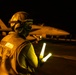 A flight deck crew member is conducting night operations on what appears to be an aircraft carrier flight deck. The individual is holding illuminated wands (light batons) used for directing aircraft during low-visibility conditions. A military jet aircraft, consistent with an F/A-18 Hornet or Super Hornet, is visible in the background.