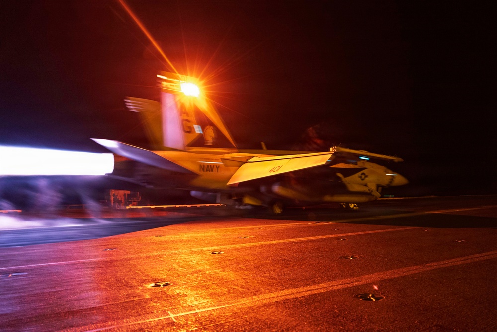 A U.S. Navy jet aircraft, marked with bureau number 401 and tail code 'AC', is captured during a night catapult launch from an aircraft carrier flight deck. The image shows significant motion blur and intense orange lighting from deck lighting, with engine exhaust visible on the left side of the frame. The aircraft appears to be an F/A-18 Hornet or Super Hornet based on its visible silhouette and markings.