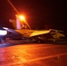 A U.S. Navy jet aircraft, marked with bureau number 401 and tail code 'AC', is captured during a night catapult launch from an aircraft carrier flight deck. The image shows significant motion blur and intense orange lighting from deck lighting, with engine exhaust visible on the left side of the frame. The aircraft appears to be an F/A-18 Hornet or Super Hornet based on its visible silhouette and markings.