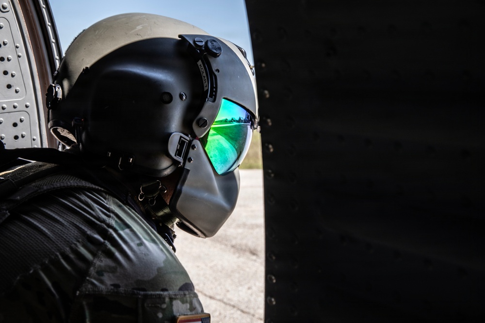 A military aviator wearing a flight helmet with a reflective green-tinted visor is photographed in close profile, positioned near the fuselage of an aircraft. The helmet appears to be a combat aviation brigade-style helmet with ear protection cups and a flip-down visor. The visor reflects what appears to be a runway or airfield environment.