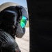 A military aviator wearing a flight helmet with a reflective green-tinted visor is photographed in close profile, positioned near the fuselage of an aircraft. The helmet appears to be a combat aviation brigade-style helmet with ear protection cups and a flip-down visor. The visor reflects what appears to be a runway or airfield environment.