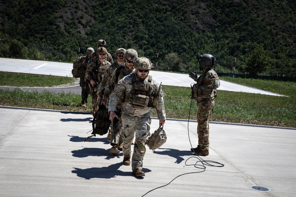A group of military personnel in full combat gear are walking in a single-file line across a concrete surface, likely a helipad or airstrip. One individual on the right, wearing a flight helmet, appears to be directing or guiding the group. The setting includes a mountainous, tree-covered backdrop under clear daylight conditions.
