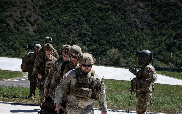 A group of military personnel in full combat gear are walking in a single-file line across a concrete surface, likely a helipad or airstrip. One individual on the right, wearing a flight helmet, appears to be directing or guiding the group. The setting includes a mountainous, tree-covered backdrop under clear daylight conditions.
