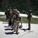 A group of military personnel in full combat gear are walking in a single-file line across a concrete surface, likely a helipad or airstrip. One individual on the right, wearing a flight helmet, appears to be directing or guiding the group. The setting includes a mountainous, tree-covered backdrop under clear daylight conditions.