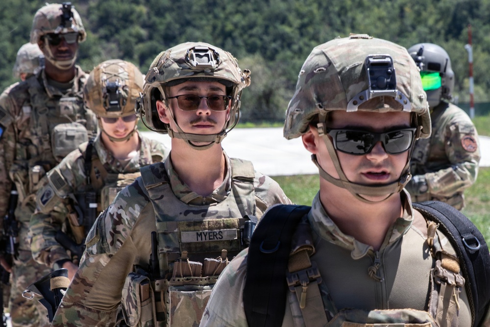 The image shows a group of military personnel in full combat gear, wearing MultiCam/OCP camouflage uniforms and tactical helmets with night vision device mounts. The soldiers are equipped with body armor, tactical vests with magazine pouches, and protective eyewear. The setting appears to be an outdoor training or operational environment with vegetation visible in the background.