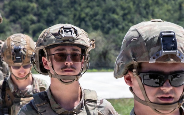 The image shows a group of military personnel in full combat gear, wearing MultiCam/OCP camouflage uniforms and tactical helmets with night vision device mounts. The soldiers are equipped with body armor, tactical vests with magazine pouches, and protective eyewear. The setting appears to be an outdoor training or operational environment with vegetation visible in the background.