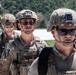 The image shows a group of military personnel in full combat gear, wearing MultiCam/OCP camouflage uniforms and tactical helmets with night vision device mounts. The soldiers are equipped with body armor, tactical vests with magazine pouches, and protective eyewear. The setting appears to be an outdoor training or operational environment with vegetation visible in the background.