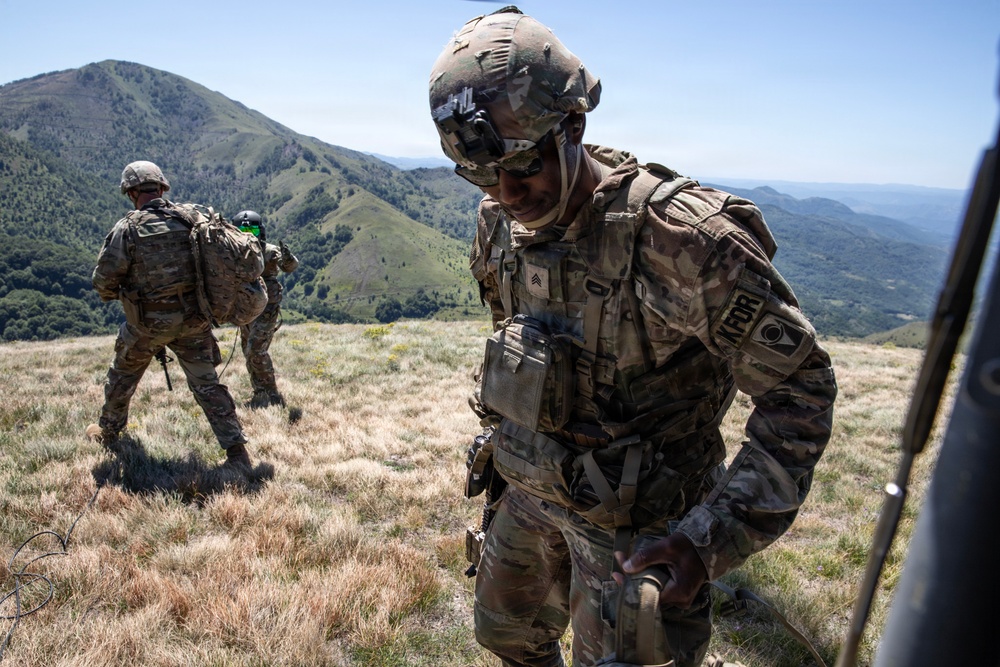 Two U.S. Army soldiers in full combat gear are operating on a mountainous hilltop under clear skies. The foreground soldier, wearing a Sergeant rank insignia and a 'KFOR' (Kosovo Force) patch, appears to be exiting or working near a vehicle. The background shows green rolling hills and a wide valley landscape.