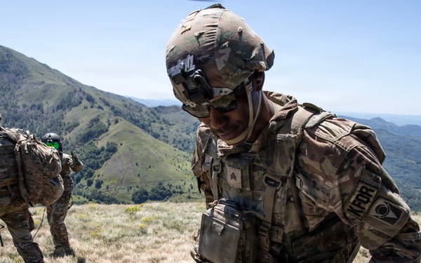 Two U.S. Army soldiers in full combat gear are operating on a mountainous hilltop under clear skies. The foreground soldier, wearing a Sergeant rank insignia and a 'KFOR' (Kosovo Force) patch, appears to be exiting or working near a vehicle. The background shows green rolling hills and a wide valley landscape.