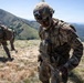 Two U.S. Army soldiers in full combat gear are operating on a mountainous hilltop under clear skies. The foreground soldier, wearing a Sergeant rank insignia and a 'KFOR' (Kosovo Force) patch, appears to be exiting or working near a vehicle. The background shows green rolling hills and a wide valley landscape.