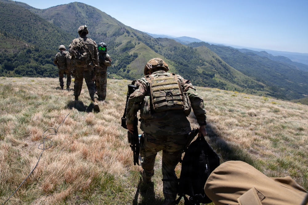 A group of military personnel in full combat gear are moving in a patrol formation across a grassy mountain ridge. They are wearing multicam/OCP camouflage uniforms and carrying tactical equipment including plate carriers and backpacks. The terrain features green, rolling mountain ranges visible in the background under clear skies.