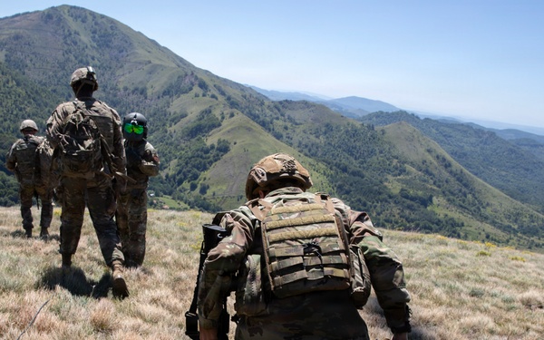 A group of military personnel in full combat gear are moving in a patrol formation across a grassy mountain ridge. They are wearing multicam/OCP camouflage uniforms and carrying tactical equipment including plate carriers and backpacks. The terrain features green, rolling mountain ranges visible in the background under clear skies.