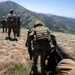 A group of military personnel in full combat gear are moving in a patrol formation across a grassy mountain ridge. They are wearing multicam/OCP camouflage uniforms and carrying tactical equipment including plate carriers and backpacks. The terrain features green, rolling mountain ranges visible in the background under clear skies.