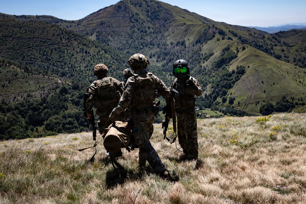 Three military personnel in full combat gear are standing on a grassy hillside overlooking a mountainous valley with dense vegetation. They appear to be observing or surveying the terrain below. One individual on the right is wearing a distinctive flight/ground crew helmet with green-tinted visor.