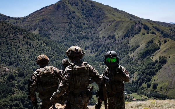 Three military personnel in full combat gear are standing on a grassy hillside overlooking a mountainous valley with dense vegetation. They appear to be observing or surveying the terrain below. One individual on the right is wearing a distinctive flight/ground crew helmet with green-tinted visor.