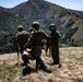 Three military personnel in full combat gear are standing on a grassy hillside overlooking a mountainous valley with dense vegetation. They appear to be observing or surveying the terrain below. One individual on the right is wearing a distinctive flight/ground crew helmet with green-tinted visor.