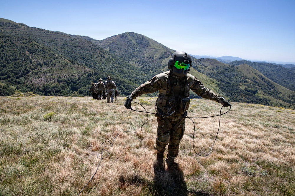 A group of military personnel in full combat gear are traversing a mountainous, grassy ridge under clear blue skies. The foreground soldier is wearing a helmet with green reflective visor and holding rope/cord lines in both hands. Additional soldiers are visible ahead in the distance moving along the ridgeline.