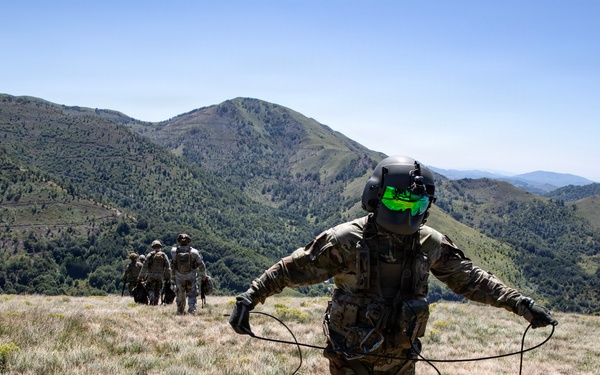 A group of military personnel in full combat gear are traversing a mountainous, grassy ridge under clear blue skies. The foreground soldier is wearing a helmet with green reflective visor and holding rope/cord lines in both hands. Additional soldiers are visible ahead in the distance moving along the ridgeline.