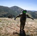 A group of military personnel in full combat gear are traversing a mountainous, grassy ridge under clear blue skies. The foreground soldier is wearing a helmet with green reflective visor and holding rope/cord lines in both hands. Additional soldiers are visible ahead in the distance moving along the ridgeline.