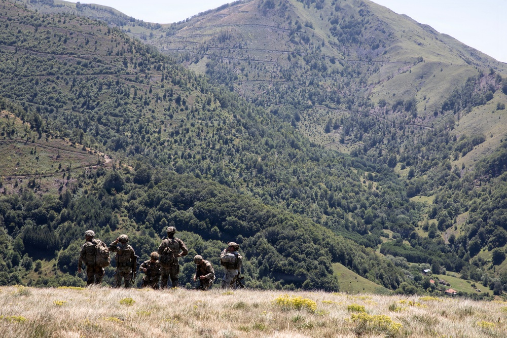 A group of approximately five soldiers in camouflage uniforms and tactical gear are positioned on a hilltop ridge overlooking a densely forested mountain valley. They appear to be observing or surveying the terrain below, with a large green mountain range serving as the backdrop. The scene suggests a military patrol or reconnaissance operation in a mountainous, rural environment.