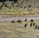 An aerial or elevated view shows a small group of approximately five individuals in military or tactical gear moving across an open grassy hillside terrain. They appear to be conducting a patrol or field exercise in a mountainous environment with scattered yellow flowering shrubs and sparse trees. The image is taken from a significant distance, suggesting observation from a high vantage point.