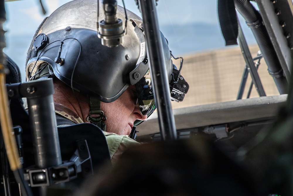 A military aviator or helicopter pilot is seated in a cockpit, wearing a flight helmet equipped with a night vision goggle mount and communications headset. The image is taken from a close, low angle inside or adjacent to the aircraft, revealing cockpit structural elements and equipment. The pilot appears to be focused downward, possibly reviewing instruments or pre-flight checks.