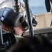 A military aviator or helicopter pilot is seated in a cockpit, wearing a flight helmet equipped with a night vision goggle mount and communications headset. The image is taken from a close, low angle inside or adjacent to the aircraft, revealing cockpit structural elements and equipment. The pilot appears to be focused downward, possibly reviewing instruments or pre-flight checks.