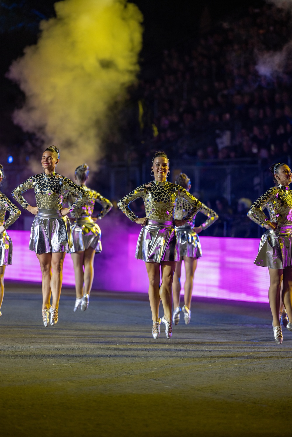 A group of female performers are executing a synchronized jump during a large-scale nighttime performance or halftime show. They are wearing matching metallic gold-and-black patterned bodysuits with silver skirts and silver lace-up heels. The venue appears to be a stadium filled with spectators, with dramatic yellow smoke and pink/purple lighting effects enhancing the spectacle.